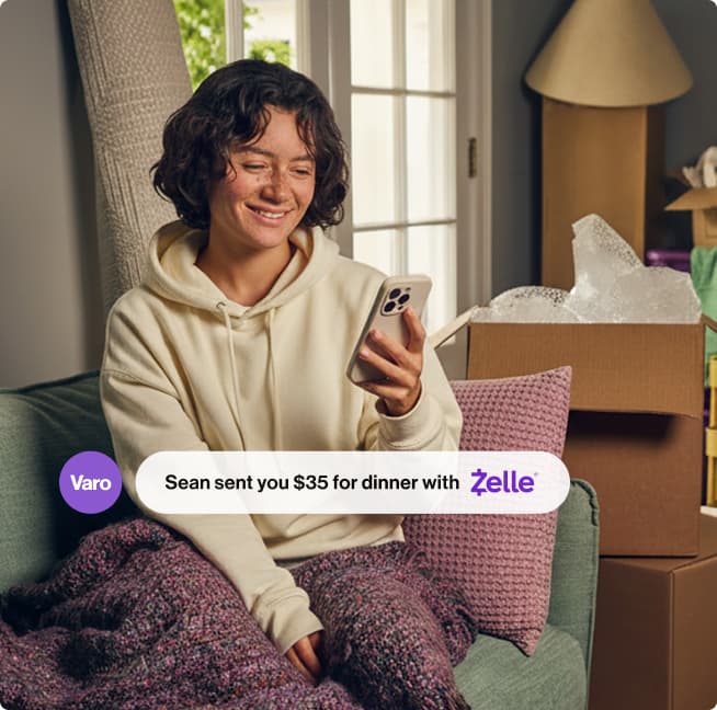 woman in cream hoodie and blanket checking phone on couch to see a payment received from Zelle in her Varo app mobile